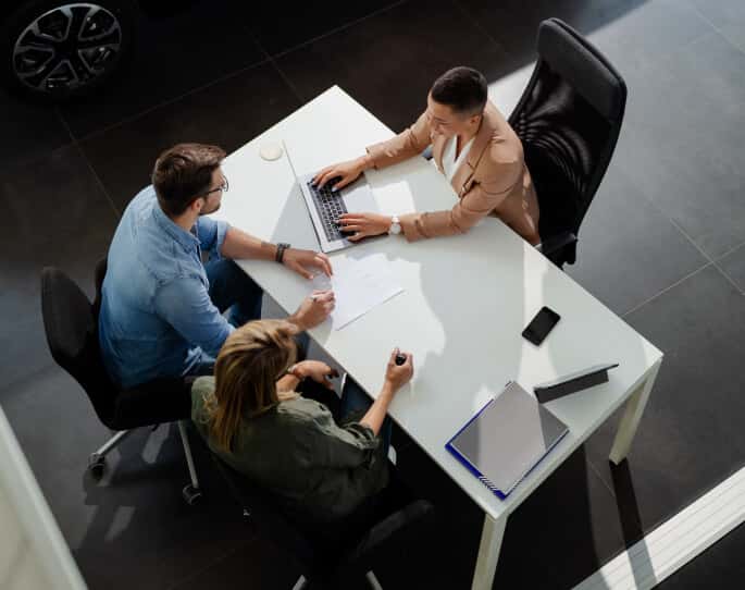 Overhead view of a couple discussing paperwork with a sales representative at a desk inside a car dealership.