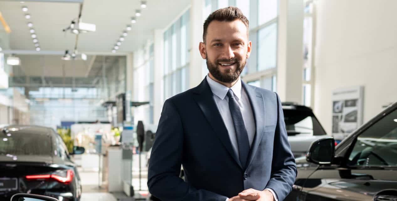 A man in a suit smiles at the camera while standing in a sunlit auto dealership.