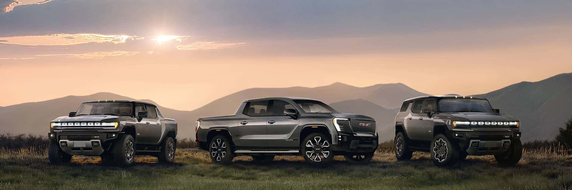 2026 EV Family exterior images with models HUMMER EV Pickup 3X and HUMMER EV SUV 3X in Meteorite Metallic color and Sierra EV Denali in Thunderstorm Gray color in country/rural, mountains and dawn environment