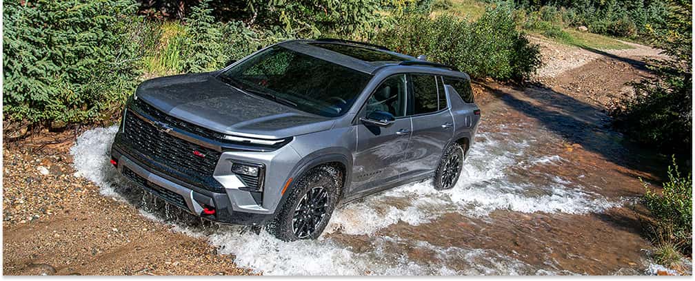 Front three-quarter view of a dark gray 2026 Chevrolet Traverse driving through a shallow stream.