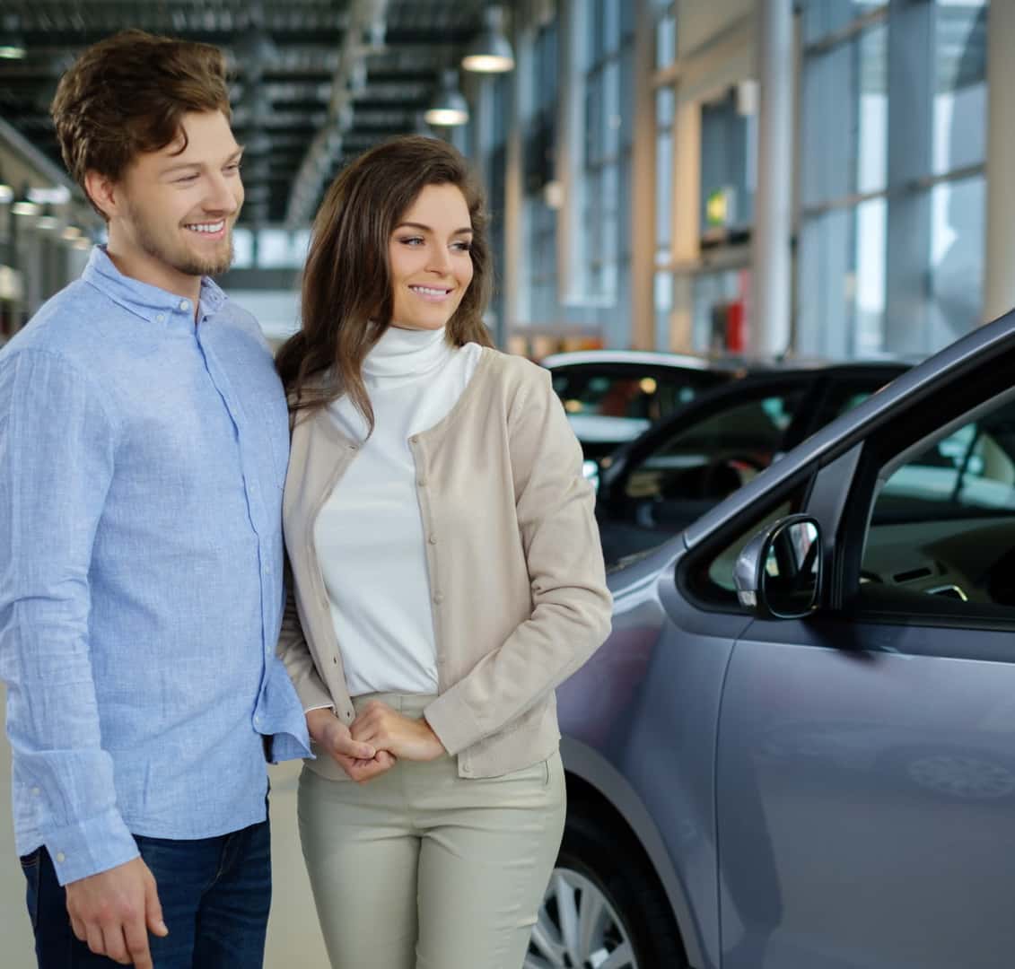 A smiling couple stands in a car showroom, looking at a silver vehicle.
