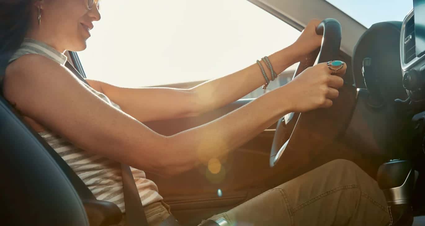 A woman wearing sunglasses and bracelets drives a car, smiling in the sunlight.

    