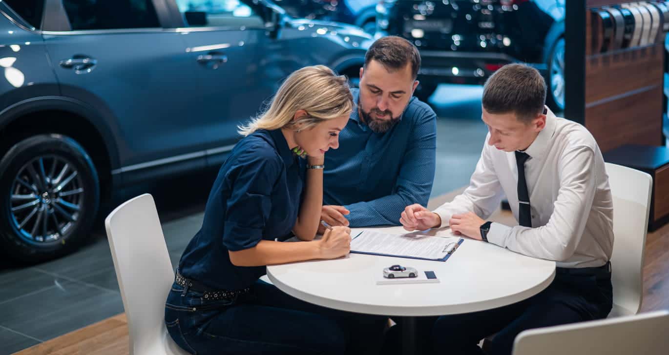 Three people sit at a round table in a car dealership, focused on signing a document.