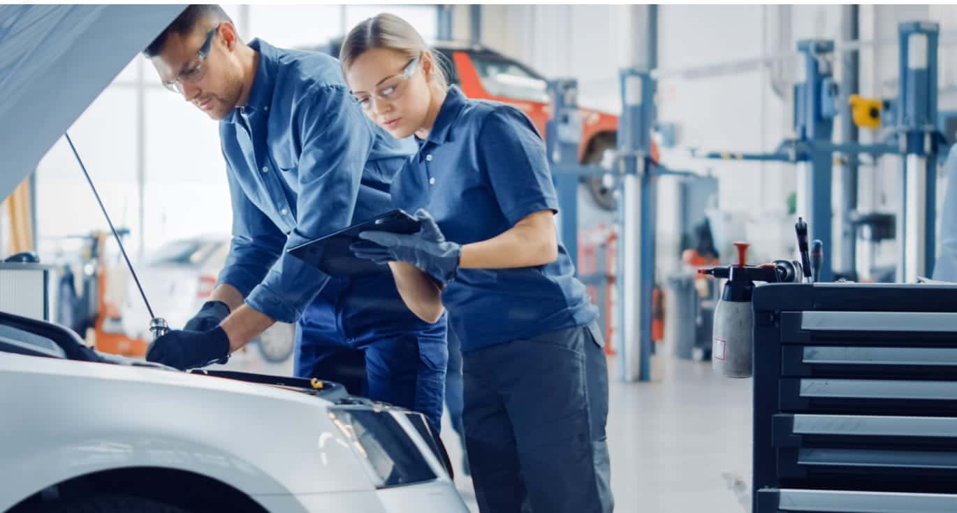 Two mechanics in a garage work on a car. Both wear safety glasses and blue uniforms.