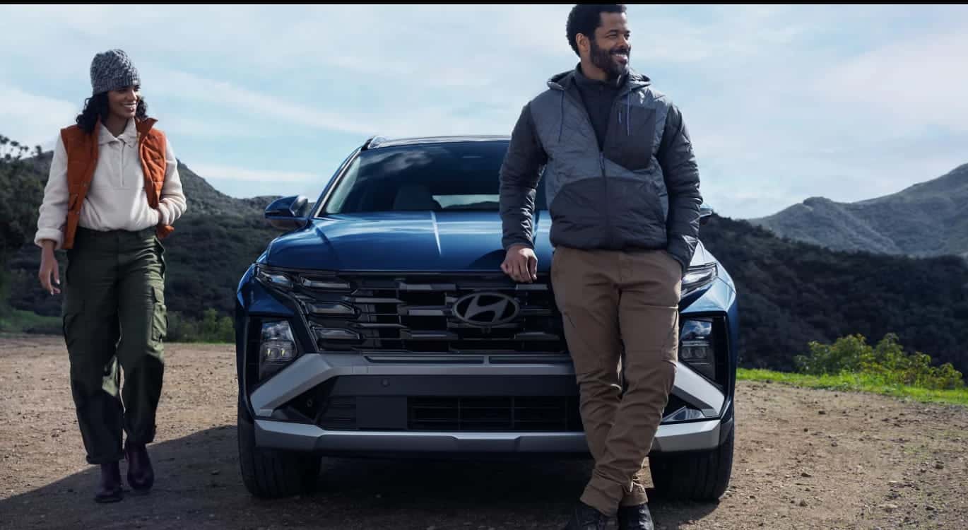 Two people standing beside a blue Hyundai Tucson in a mountainous outdoor setting.