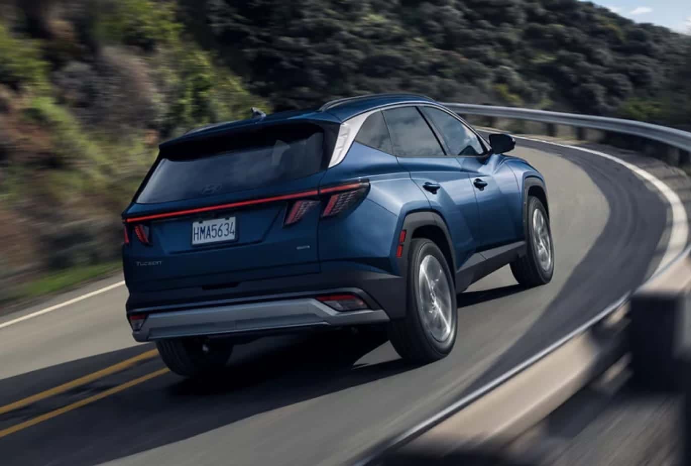 Blue Hyundai Tucson driving on a winding road, shown from the rear three-quarter view.