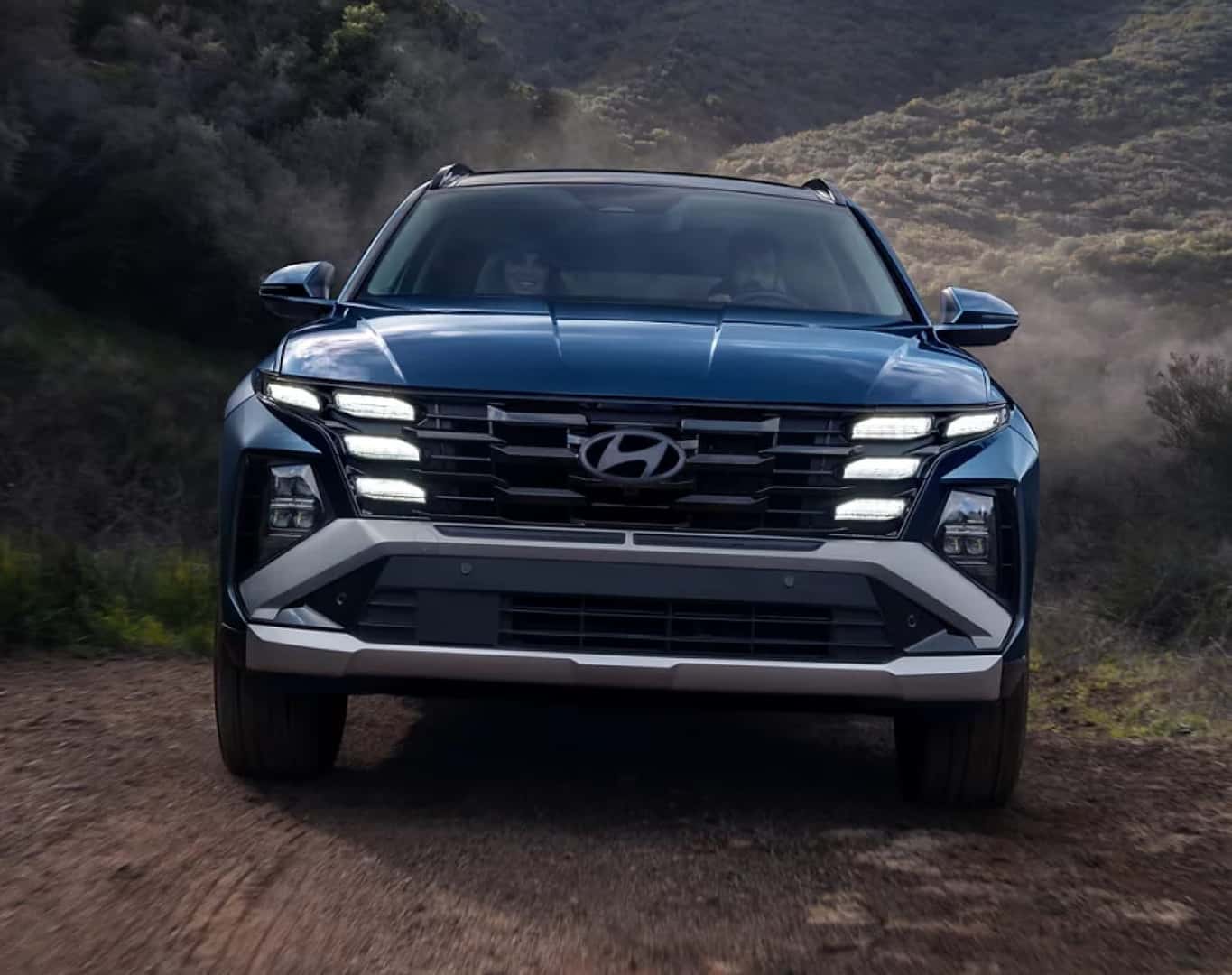 Blue Hyundai Tucson driving on a rugged mountain road, shown from the front with its LED headlights illuminated.