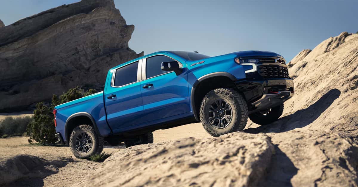 Blue Chevy Silverado 1500 pickup truck driving over rocky terrain in a desert landscape.