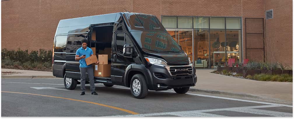 Black 2026 RAM ProMaster high-roof cargo van with open side door parked outside a modern brick building.