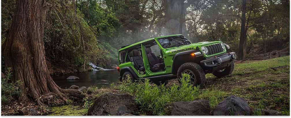 Green 2026 Jeep Wrangler Rubicon with doors removed parked near a stream in a forest.