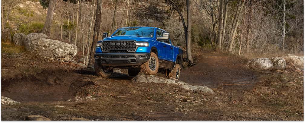 Blue 2026 RAM 1500 Rebel pickup truck driving over rocky terrain on a dirt trail.