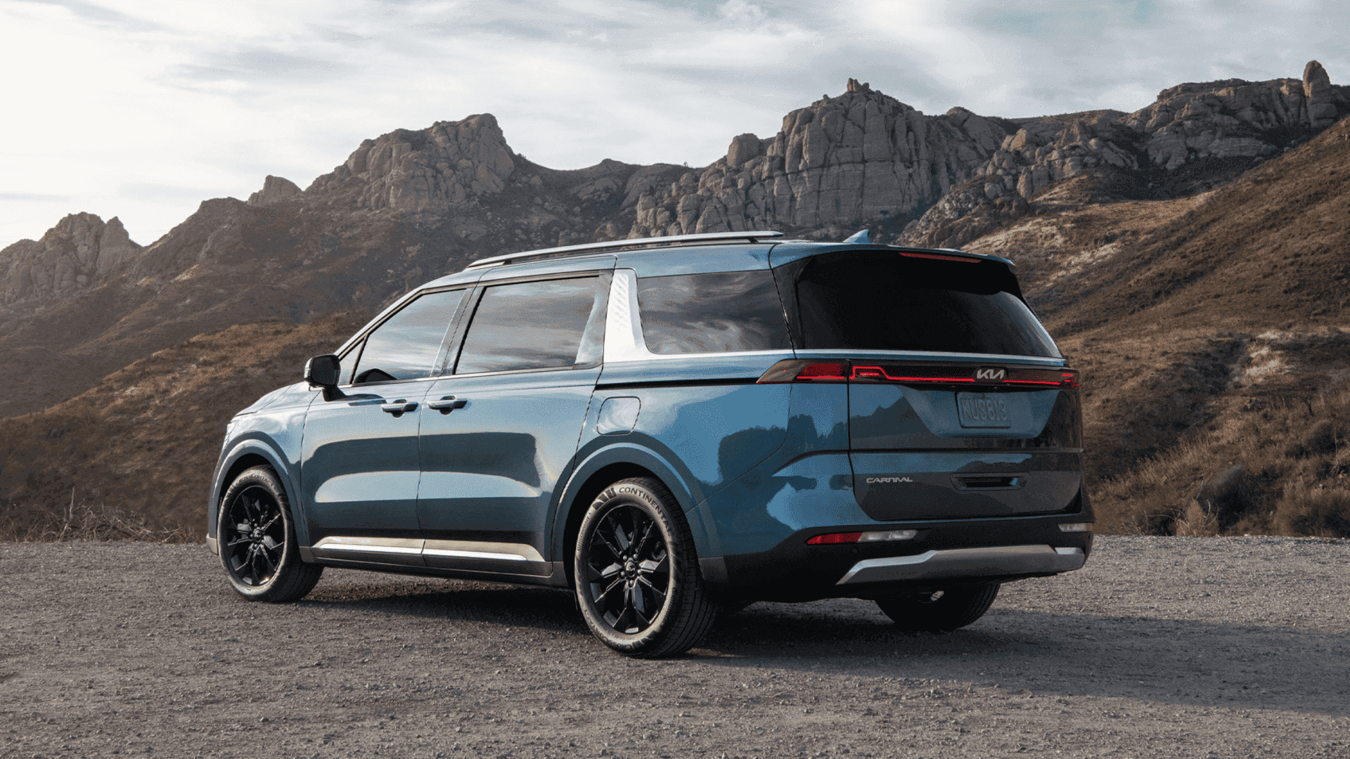 Rear view of blue Kia Carnival parked near rocky hills under cloudy sky