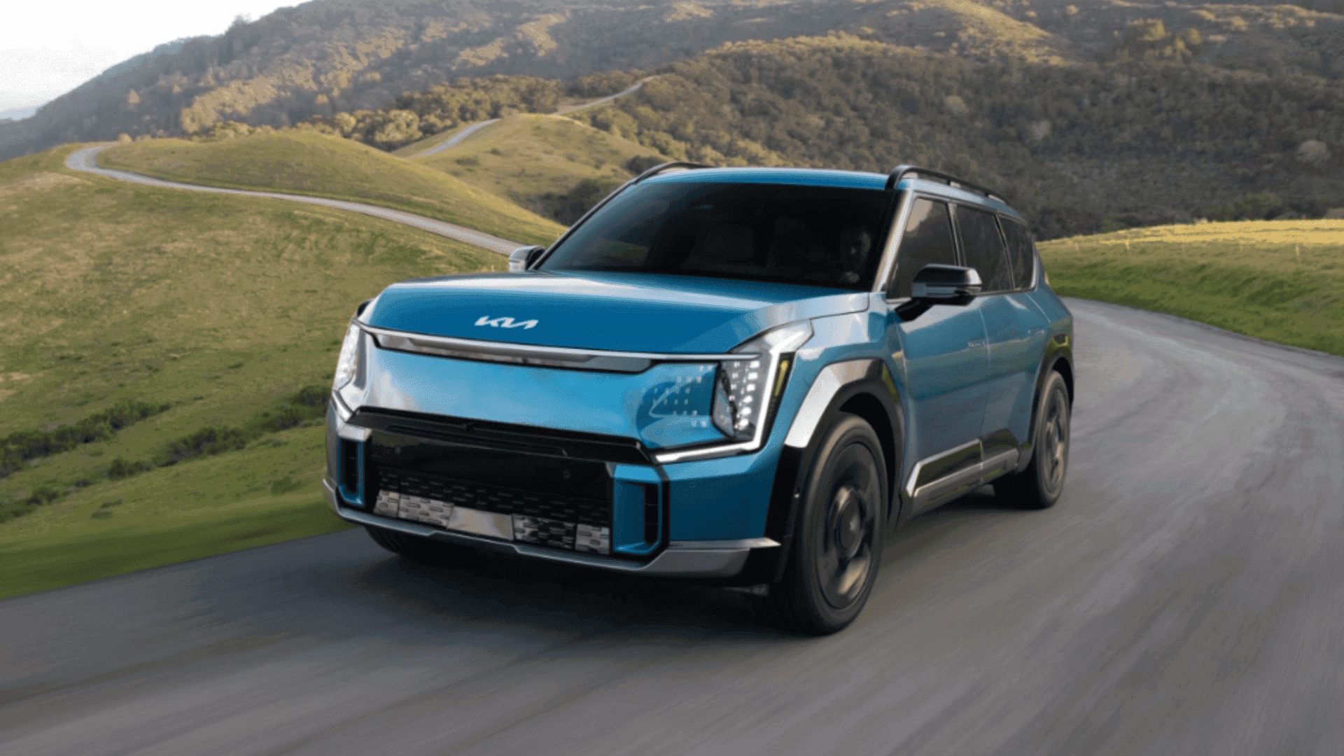 Blue 2024 Kia EV9 navigating a scenic, winding road through grassy hill