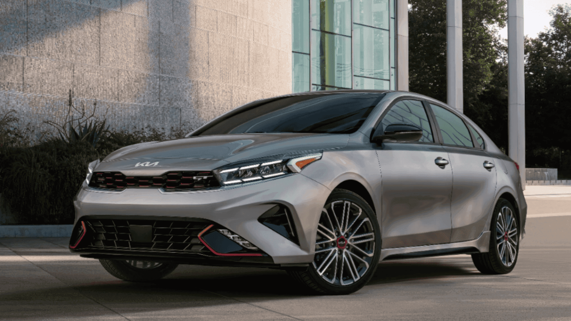 Silver 2024 Kia Forte GT sedan parked in front of a modern glass building.