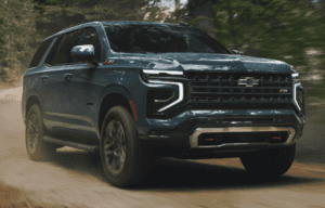 A 2025 Chevrolet Tahoe Z71 in metallic blue powers through a dirt trail in a forest, kicking up dust. The SUV features off-road styling with a bold black grille, red tow hooks, and rugged tires built for adventure.