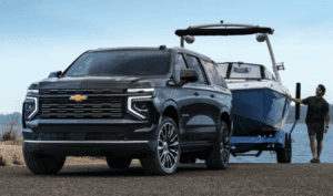 A 2025 Chevrolet Suburban SUV in dark gray is parked on a boat ramp beside a lake, towing a sleek blue and white wake boat. A man stands beside the trailer, preparing for launch under a clear blue sky.