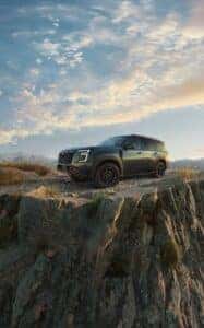 2025 Nissan Armada parked dramatically on a rocky cliff edge at sunset, highlighting its bold new front grille, rugged tires, and off-road capability with a scenic mountain backdrop and vibrant sky.