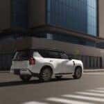 Rear view of a white 2025 Nissan Armada driving through a modern urban intersection, showcasing its bold new design, black roof, and LED tail lights against a backdrop of glass office buildings.