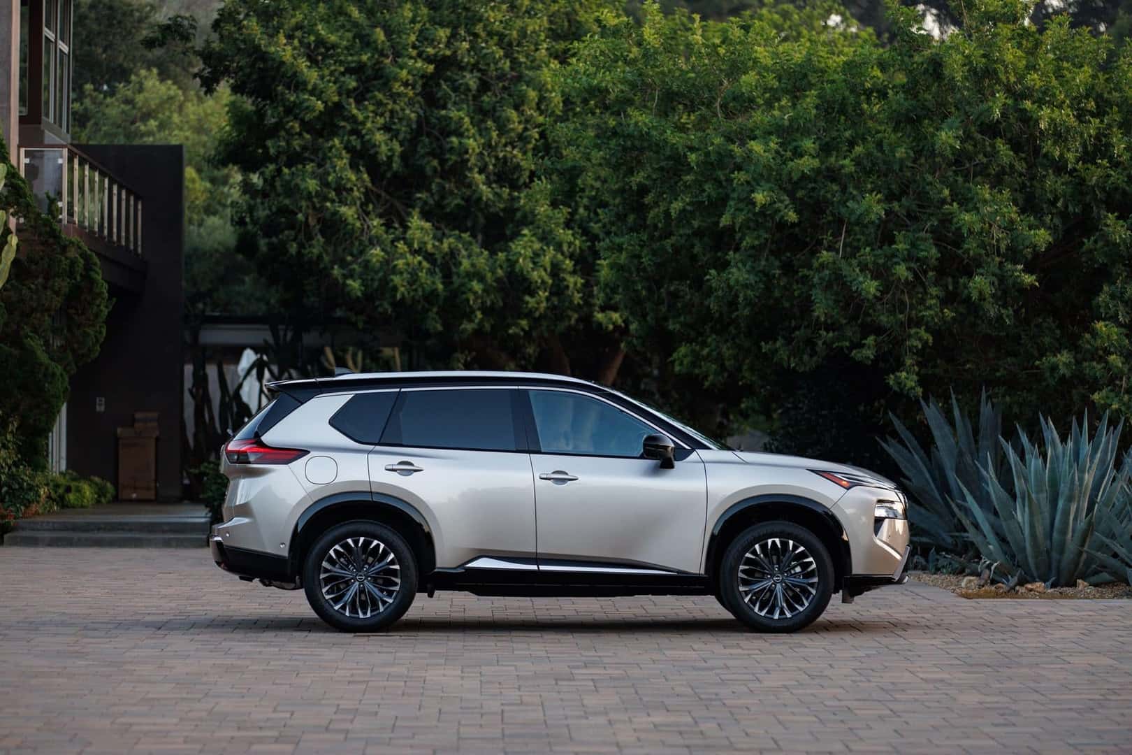 The Shimmering Side Profile Of The Family Friendly Nissan Rogue, Parked Near A Recreational Park Of Green Trees