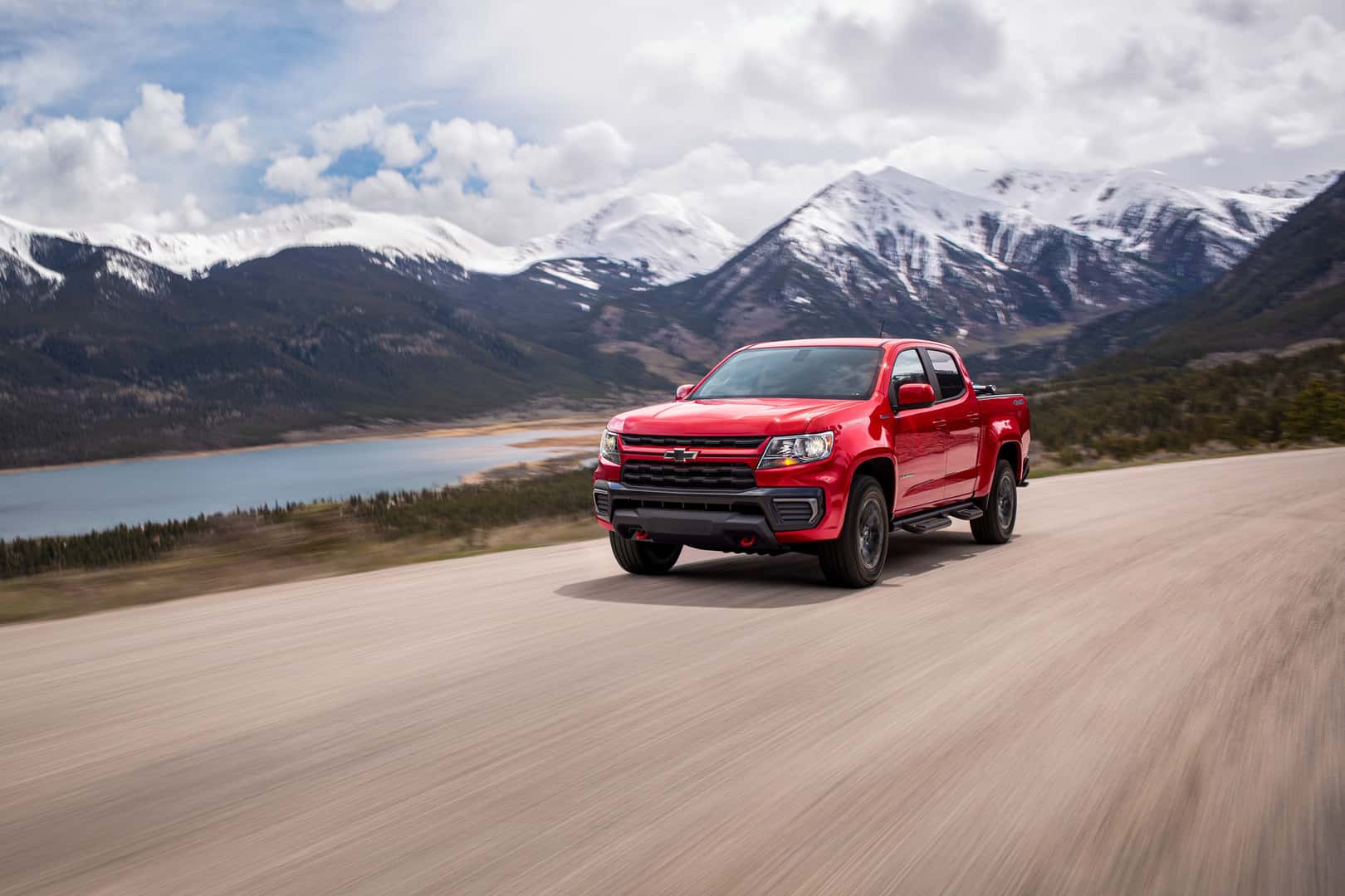 A red used 2022 Chevy Colorado in Phoenix drives towards the viewer to the left on a dirt road with a lake and snowcapped mountains in the background.