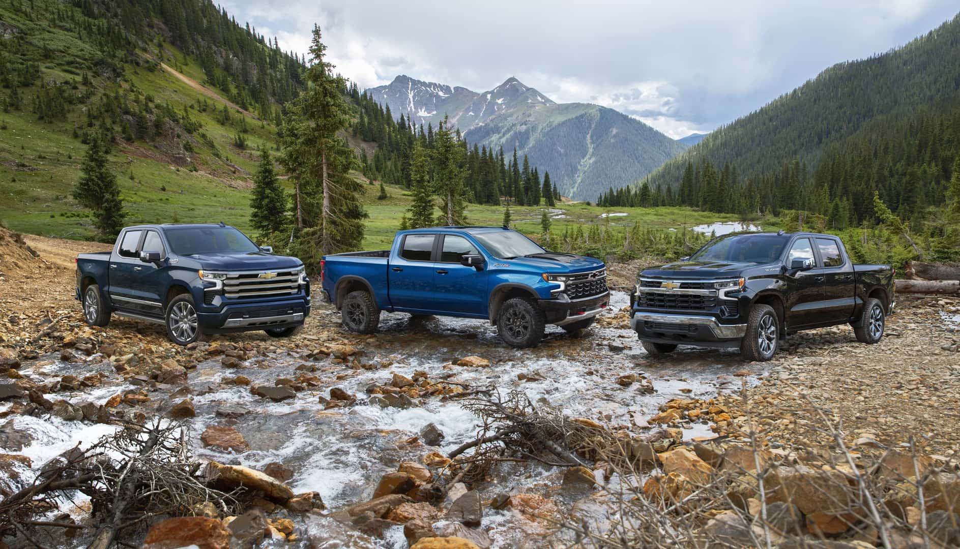 Three used 2023 Chevy Silverado 1500s in Phoenix are parked on a stream in a remote mountain valley.