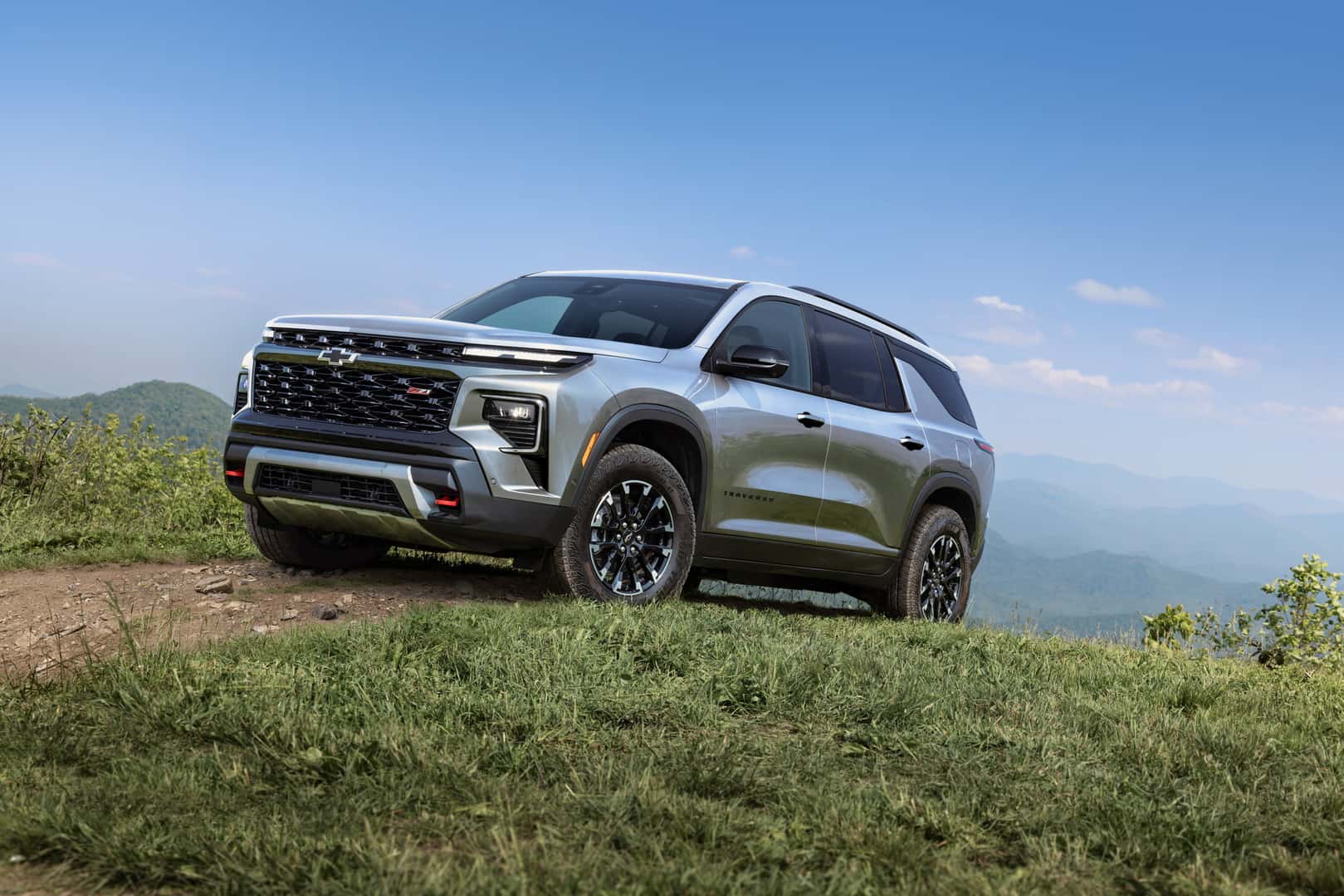 A silver used 2024 Chevy Traverse in Phoenix drives towards the viewer to the left up a dirt trail at the top of an alpine grassy hill.