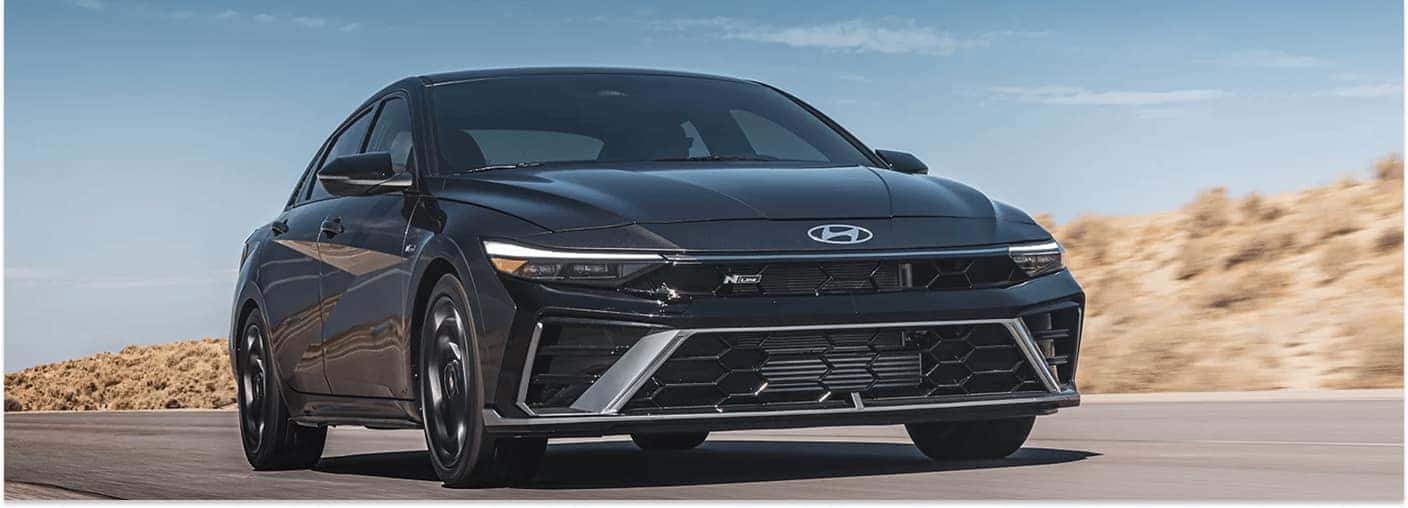 A black 2026 Hyundai Elantra N Line sedan drives along an open road, set against a backdrop of arid hills and a clear blue sky.