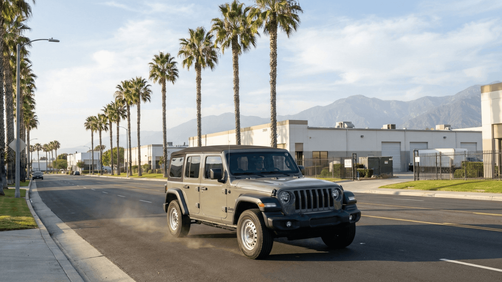 A gray four-door Jeep Wrangler driving down a sunny, palm-tree-lined street with mountains in the background.
