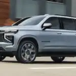 The 2026 Chevrolet Suburban near Ruth NC in silver blue with Suburban badging, bold Bowtie grille, LED signature lighting, and large multi-spoke wheels driving past a modern urban building