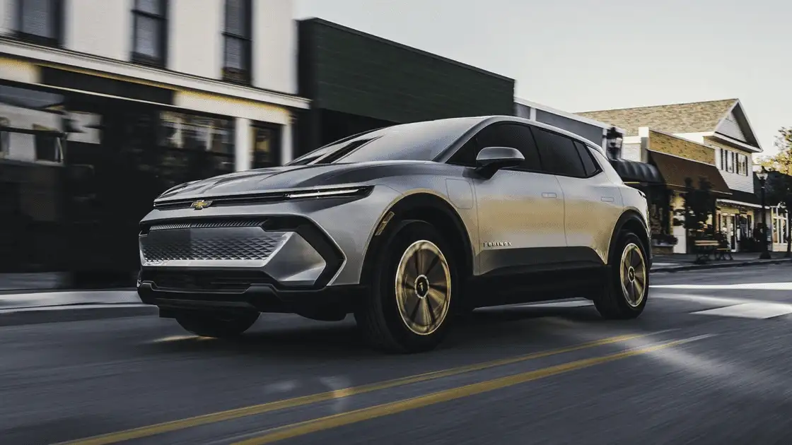 2026 Chevrolet Equinox EV near Ruth NC in silver with illuminated Bowtie grille, Equinox badging, aerodynamic profile, and gold-tone turbine wheels driving through a small town street