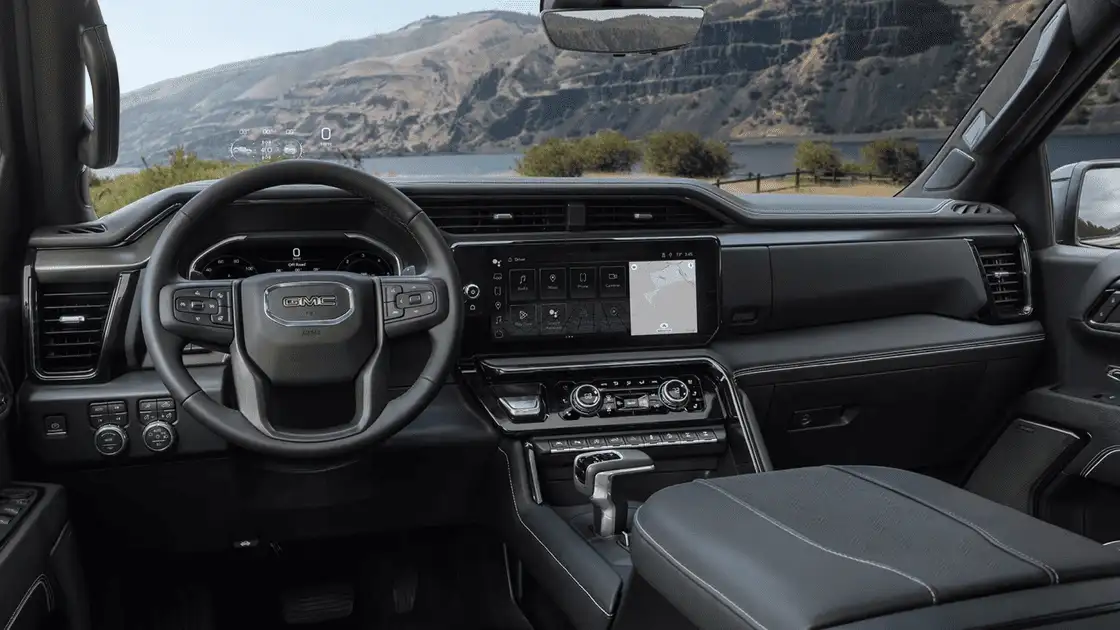 2026 GMC Sierra 1500 interior dashboard near Ruth NC with GMC-branded steering wheel, touchscreen infotainment showing Maps and Audio, heads-up display, and lakeside mountain backdrop