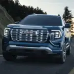 Why the 2026 GMC Terrain Denali competes well around Ruth NC in dark steel blue with chrome GMC grille lettering, LED headlights, and premium alloy wheels driving on a winding forest road