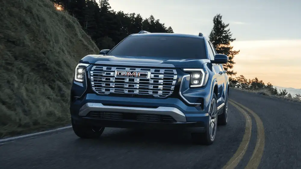 Why the 2026 GMC Terrain Denali competes well around Ruth NC in dark steel blue with chrome GMC grille lettering, LED headlights, and premium alloy wheels driving on a winding forest road