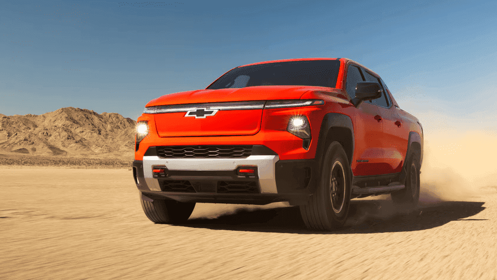 2026 Chevrolet Silverado EV near Ruth NC in orange with illuminated Bowtie grille, LED headlights, Trail Boss badging, and aggressive off-road tires driving through a desert landscape