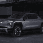 Built for more than the jobsite the 2026 Chevrolet Silverado EV near Ruth NC in dark gray with illuminated Bowtie grille, LED light bar, and multi-spoke alloy wheels plugged into a home EV charger in a modern garage