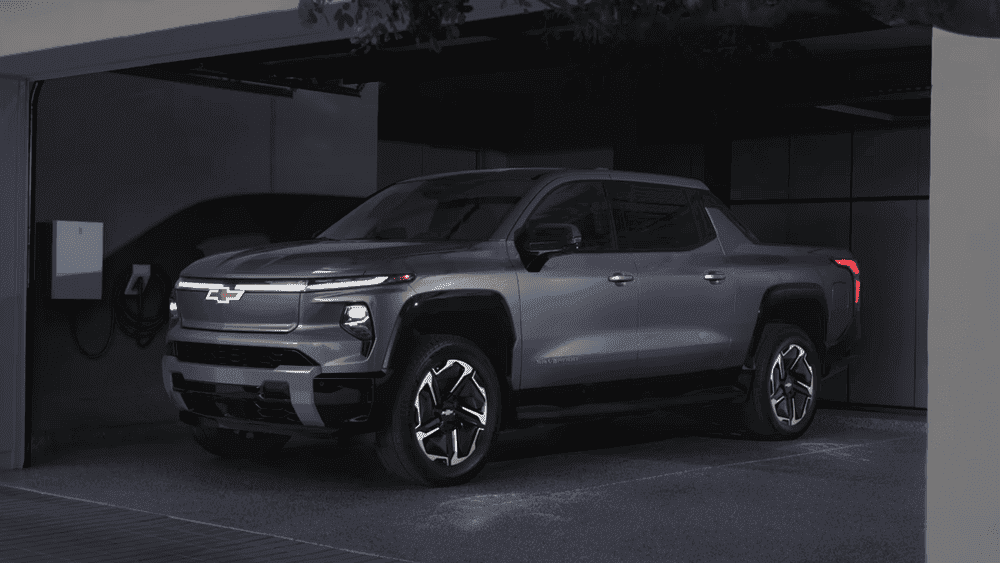 Built for more than the jobsite the 2026 Chevrolet Silverado EV near Ruth NC in dark gray with illuminated Bowtie grille, LED light bar, and multi-spoke alloy wheels plugged into a home EV charger in a modern garage