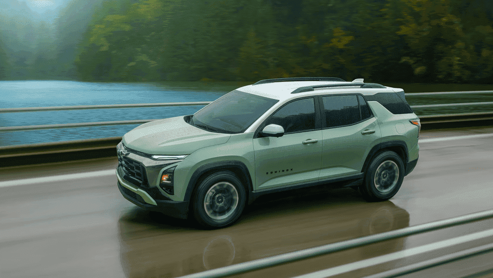 New this year the 2026 Chevrolet Equinox near Ruth NC in sage green with white roof, Equinox badging, Bowtie grille, and dark alloy wheels driving along a lakeside highway