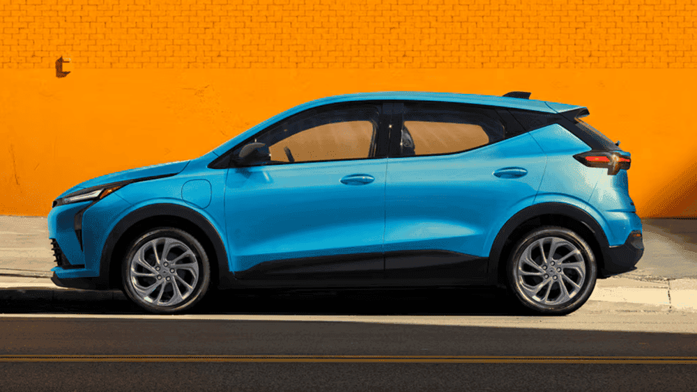 2027 Chevrolet Bolt near Ruth NC in bright blue with black lower cladding and silver alloy wheels against orange wall