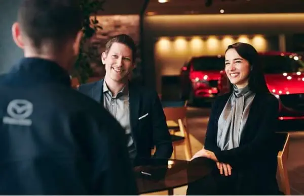 Couple is talking to Mazda Dealership Employee inside of showroom. 