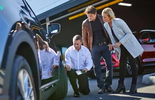 Couple and Mazda Service Tech are inspecting front tire of vehicle outside the dealership. 