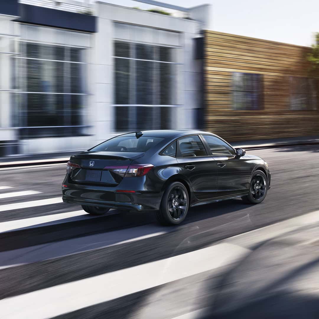 A dark-colored Honda Civic Sedan driving down a city street past modern buildings.