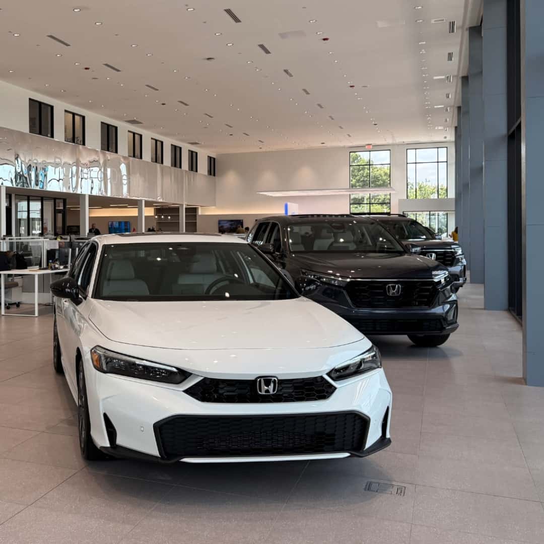 A white Honda Civic sedan displayed inside the Greensboro Honda dealership showroom, with a dark Honda CR-V parked just behind it.