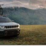 Silver 2026 Jeep Cherokee with a roof-mounted bicycle parked in a field, front three-quarter view.