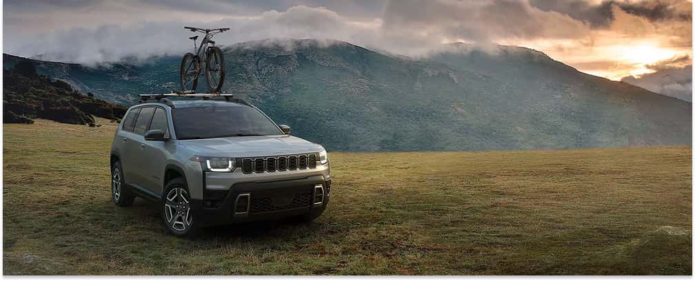 Silver 2026 Jeep Cherokee with a roof-mounted bicycle parked in a field, front three-quarter view.