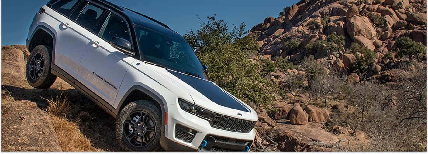 A white 2026 Jeep Grand Cherokee with a black roof and blue front tow hooks climbs a steep, rocky incline under a clear blue sky.