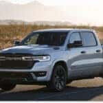 A silver 2026 Ram 1500 pickup truck with black wheels and a prominent front grille featuring the RAM logo is parked on an asphalt road in a sunlit desert landscape with mountains in the distance.