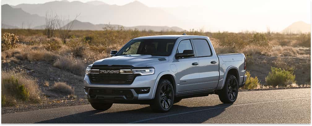 A silver 2026 Ram 1500 pickup truck with black wheels and a prominent front grille featuring the RAM logo is parked on an asphalt road in a sunlit desert landscape with mountains in the distance.