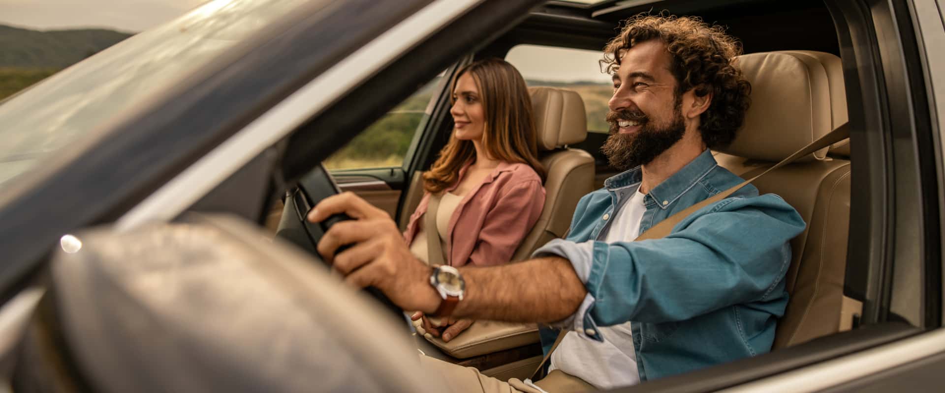 A man and woman are seated in a car, both focused on the road ahead while driving.
                    