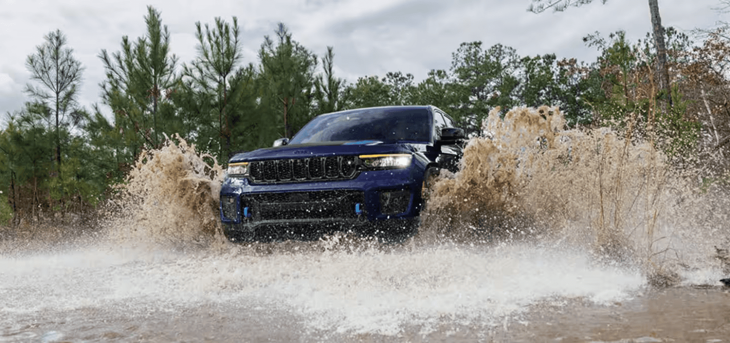 Jeep Grand Cherokee Trailhawk splashing through a muddy water trail surrounded by forest, showcasing off-road performance and rugged capability.