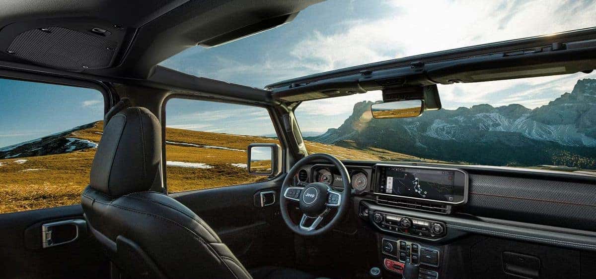 Interior view of the 2025 Jeep Wrangler with the roof removed, showcasing the steering wheel, touchscreen display, and panoramic mountain landscape.